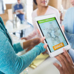 A group of unrecognizable women stand in a circle at their local food bank and hold up a digital tablet. The screen reads, "Make a Difference. Donate".