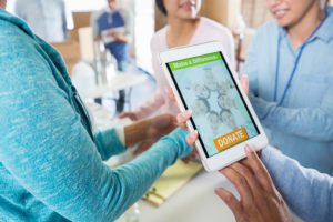 A group of unrecognizable women stand in a circle at their local food bank and hold up a digital tablet. The screen reads, "Make a Difference. Donate".