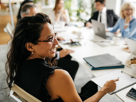 Over the shoulder profile of bespectacled female executive in early 30s sitting at conference table and laughing as she interacts with off-camera colleague.