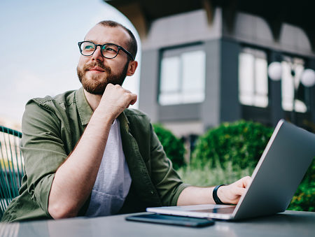 Young handsome male 20 years old in eyeglasses sitting at street cafe during free time with new modern laptop, bearded man freelancer thoughtfully looking aside and thinking about distance work